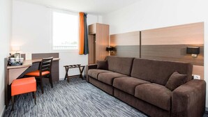 Ein modernes Hotelzimmer mit Sofa, Schreibtisch mit Stuhl, kleinem Tisch und einem Fenster mit Vorhängen.