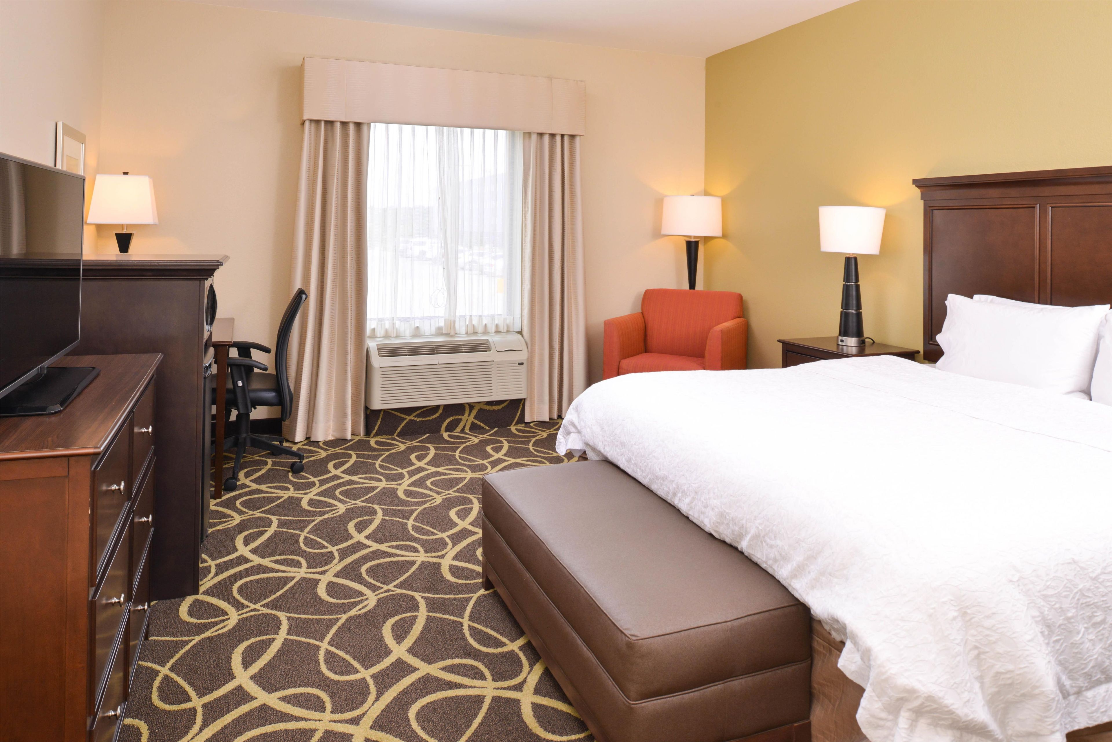 A hotel room with a bed, a desk, a chair, a bench, a lamp, and an air conditioning unit.