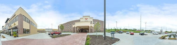 A hotel with a central entrance, a parking lot with multiple cars, and a nearby building with a sign. - Hampton Inn Omaha Midtown-Aksarben Area (Omaha)