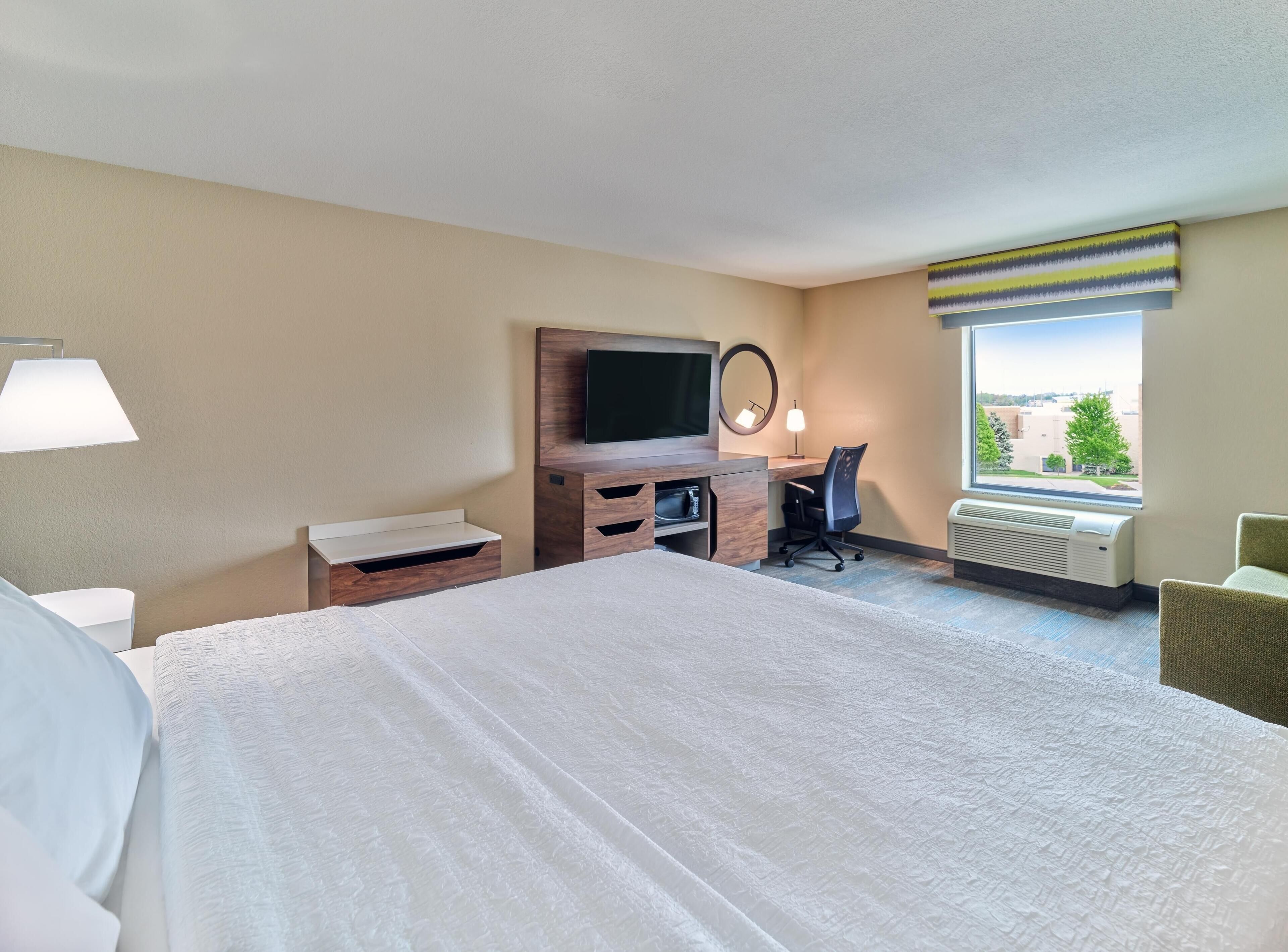 Habitación de hotel con una cama grande, un escritorio con computadora, un televisor, un espejo y una ventana con vistas.