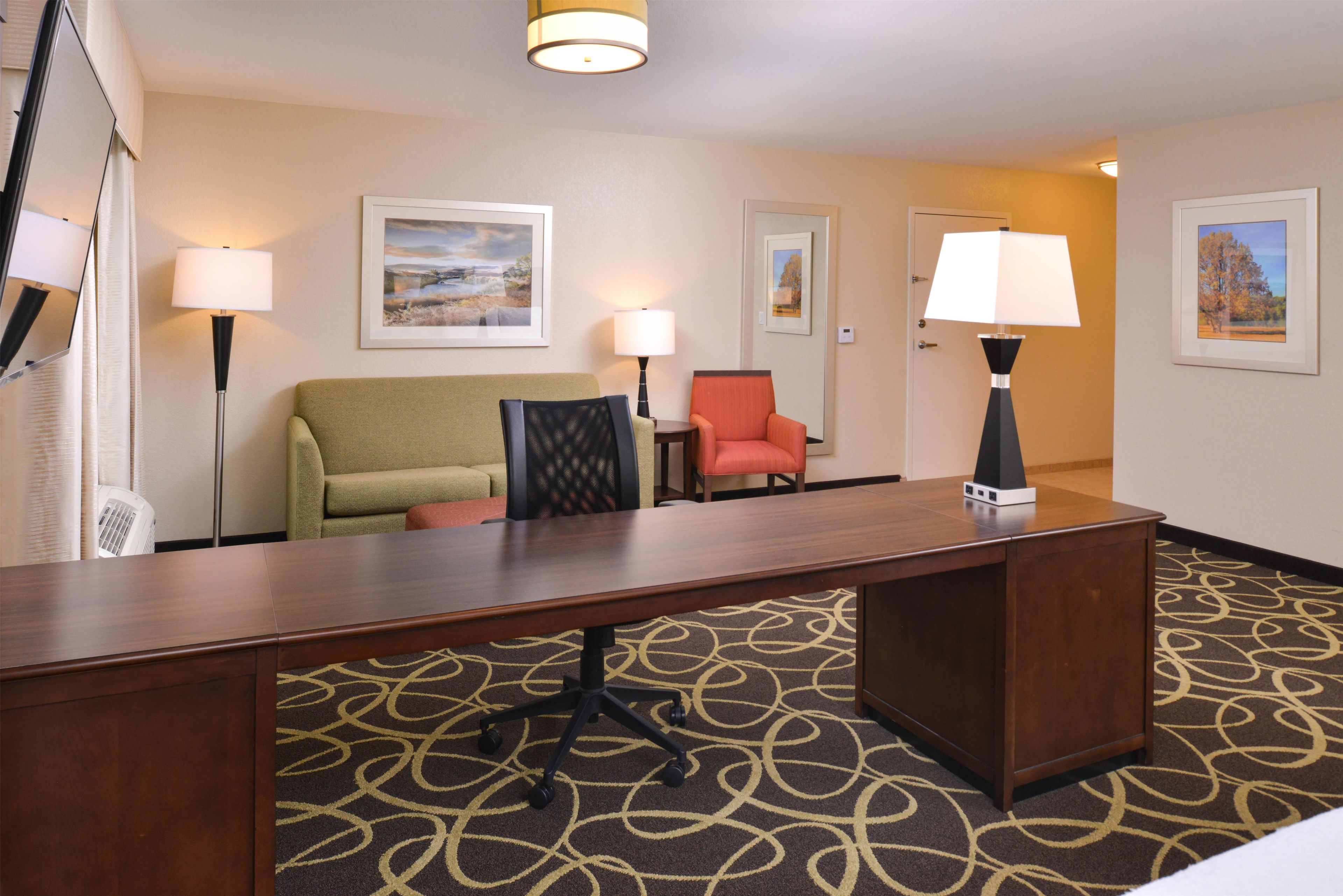 A hotel room with a wooden desk, a green sofa, a red chair, and a patterned carpet.
