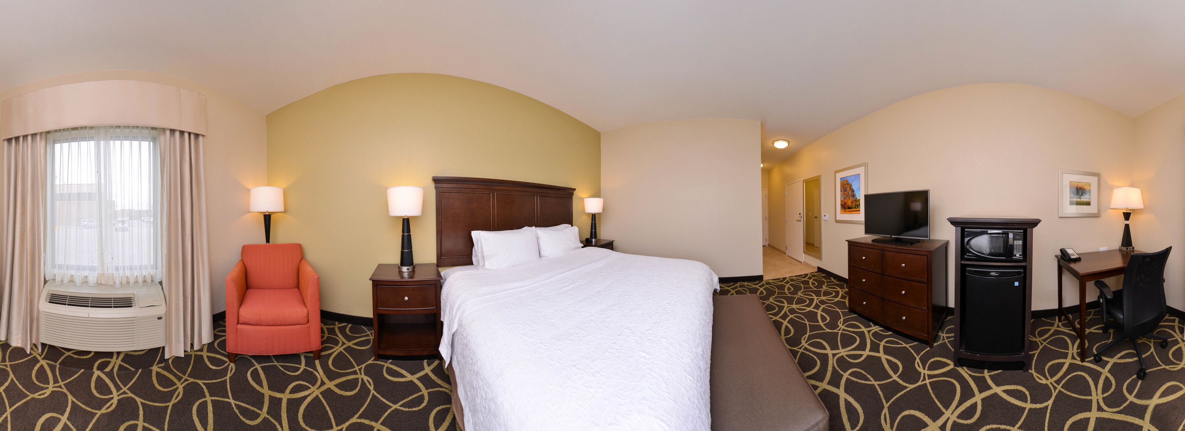 A hotel room with a large bed, a desk with a chair, a TV, a dresser, and a window with curtains.