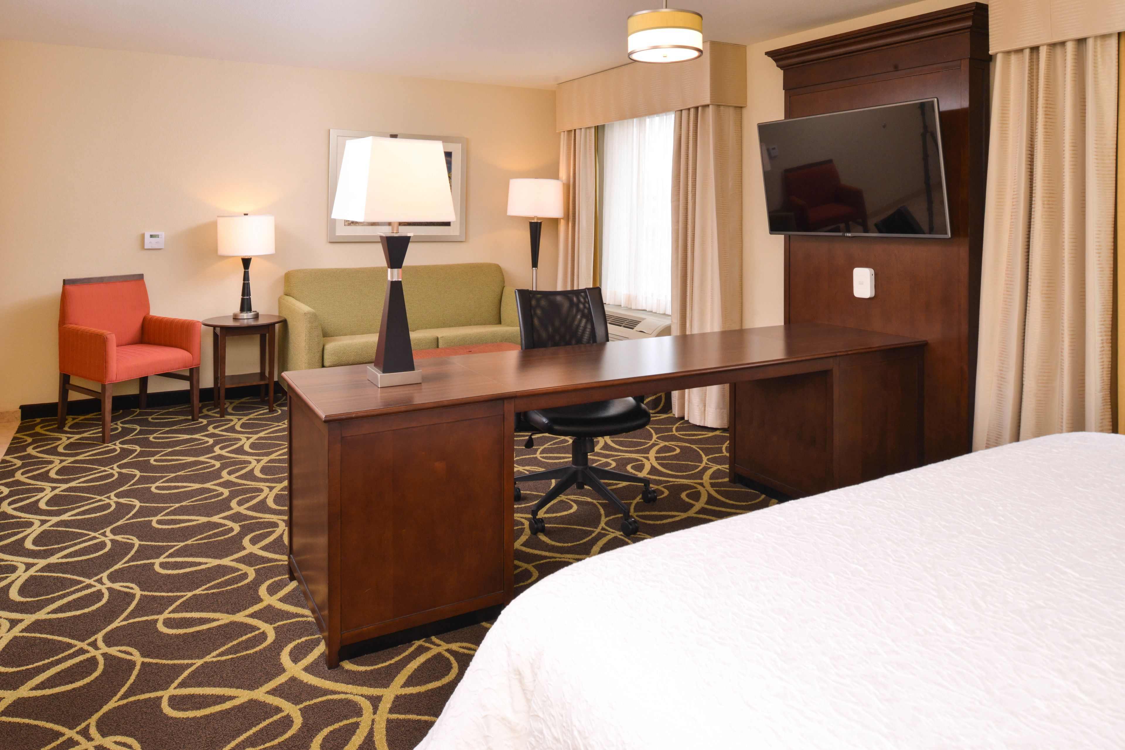 A hotel room with a bed, a desk, two chairs, a TV, and a lamp.