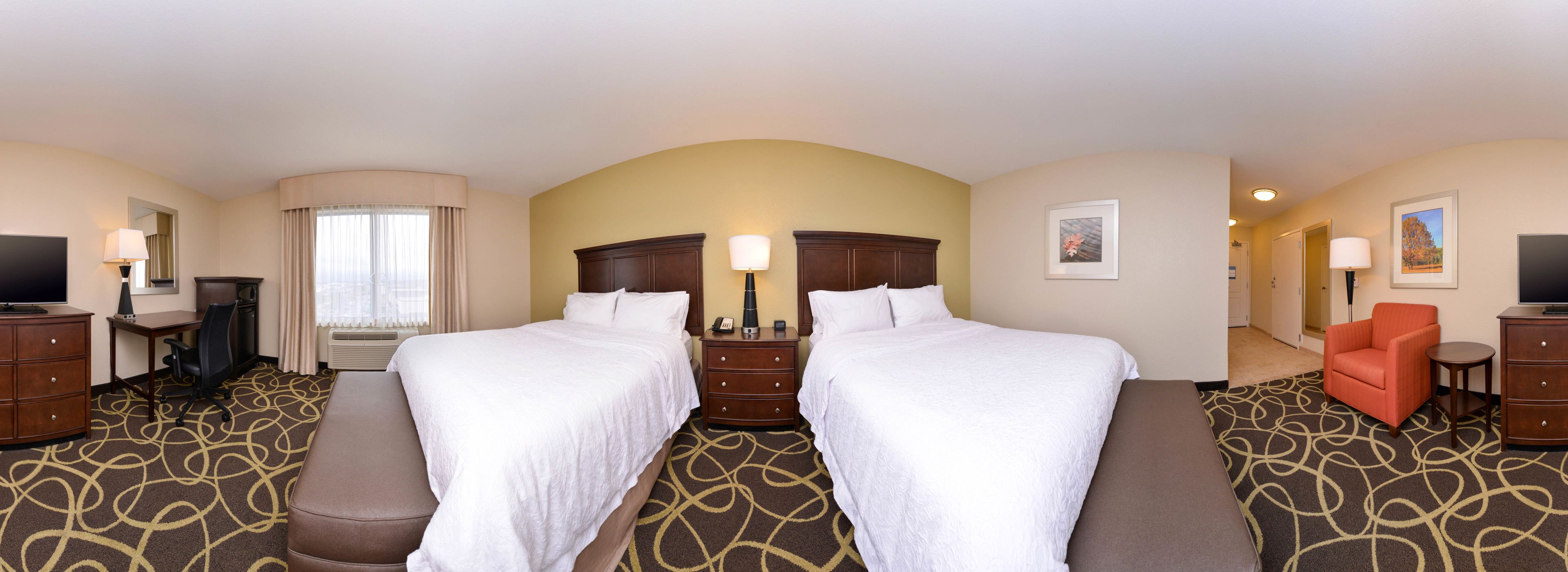 A hotel room with two beds, a desk with a chair, a TV, a dresser, and a red armchair.