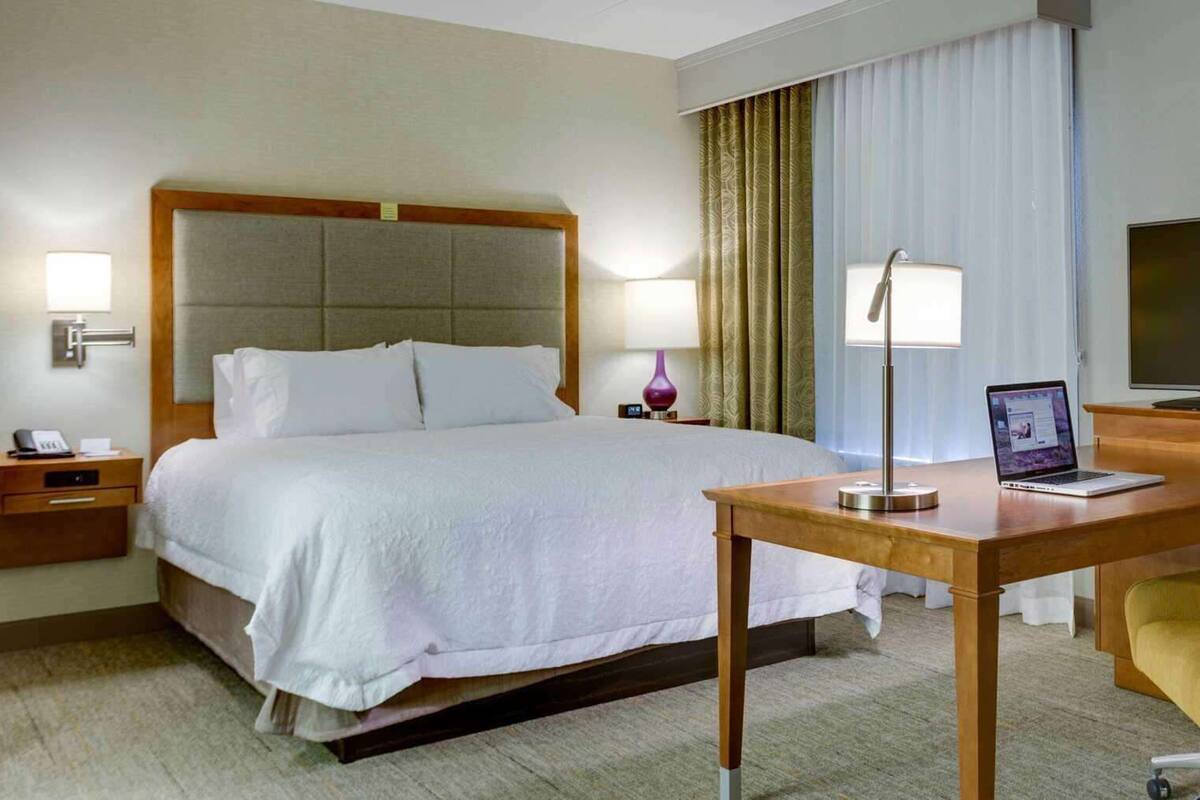 A hotel room with a bed, bedside table, lamp, desk, chair, TV, and curtains.