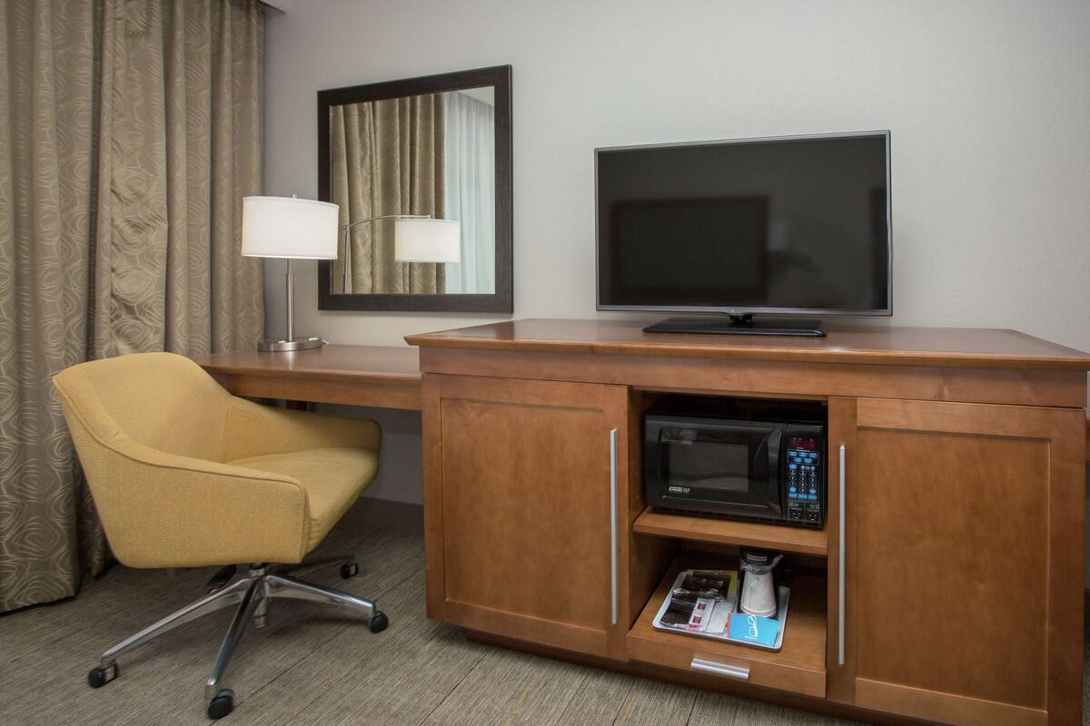 A hotel room with a wooden desk, a television, a lamp, a microwave, and a chair.