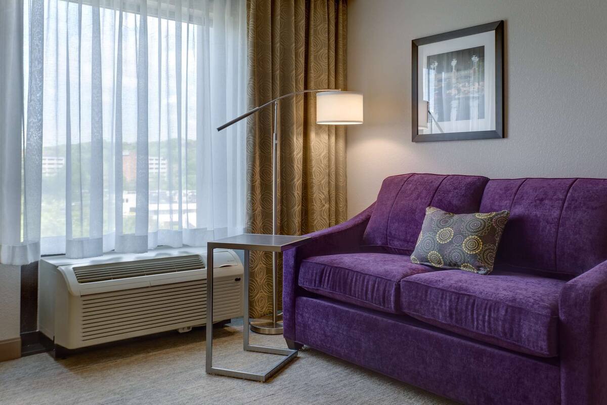 A purple sofa with a patterned pillow, a floor lamp, a framed picture on the wall, and a window with sheer curtains.