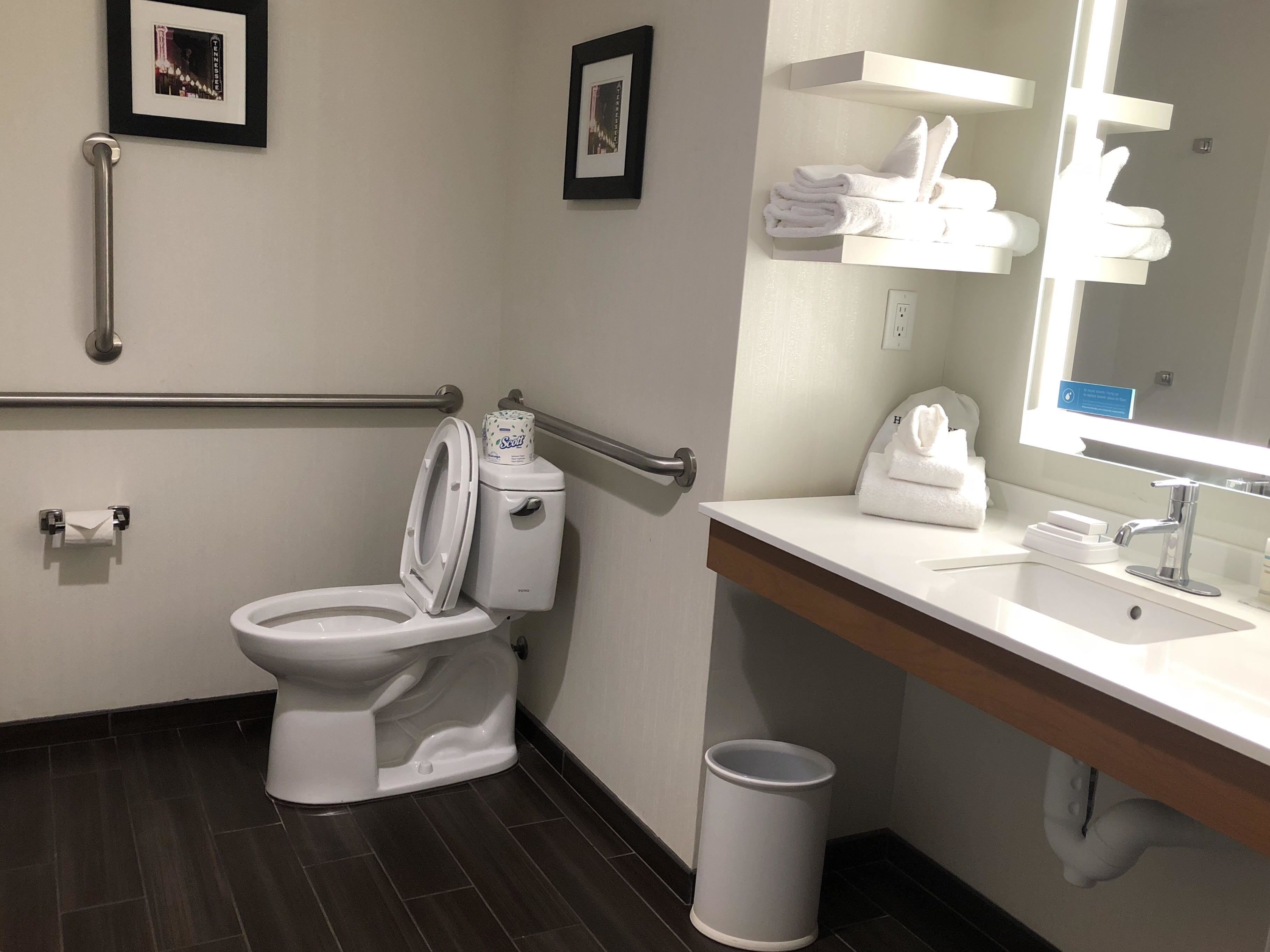Un bany modern amb vàter, baranes, un lavabo, un mirall i un cubell de basures.