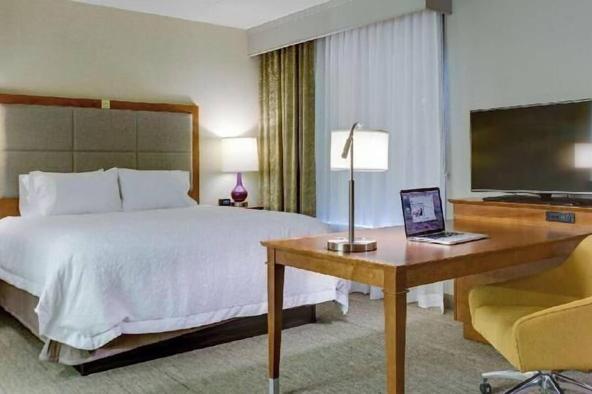A hotel room with a bed, bedside tables, a desk with a laptop, a chair, a TV, and curtains.