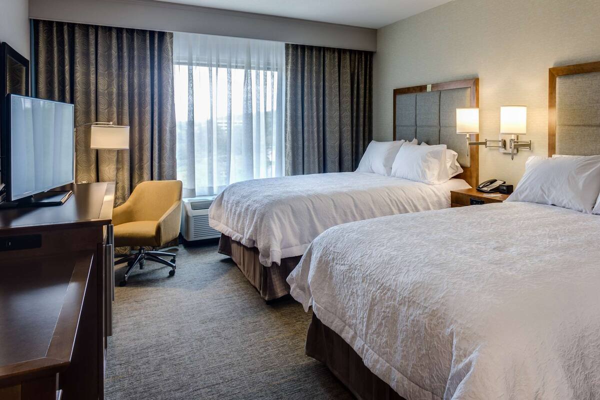 A hotel room with two beds, a desk, a chair, and a TV.