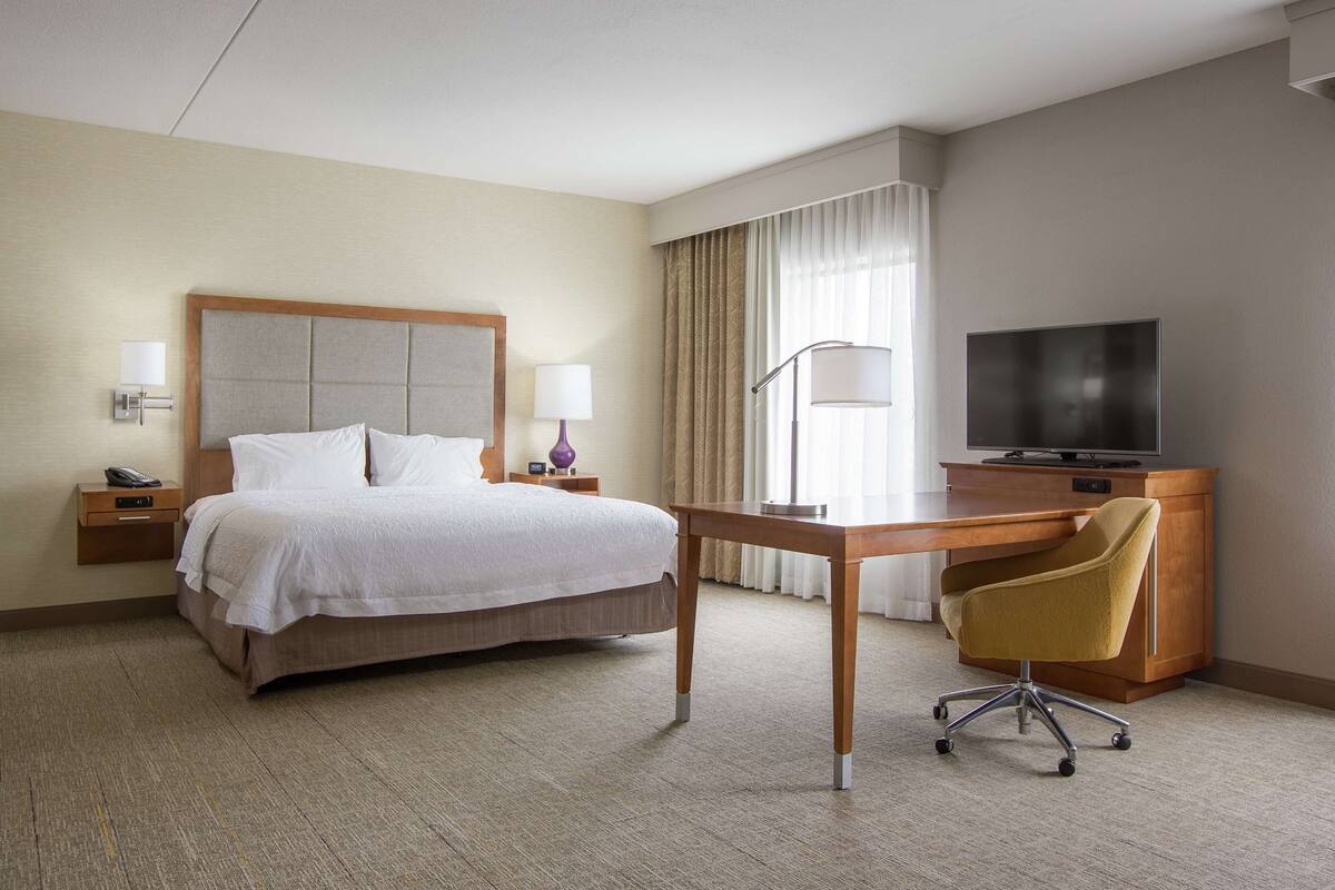 A hotel room with a bed, bedside tables, a desk, a chair, a television, and curtains.