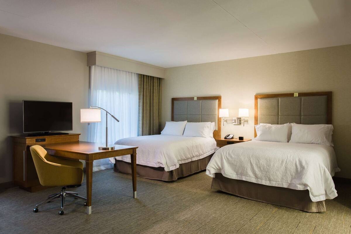 A hotel room with two beds, a desk with a TV, a chair, and a lamp.