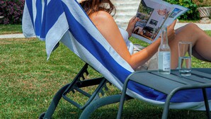 A person relaxing on a lounge chair with a book and a bottle, in front of a historic building with arched windows and a tiled roof. - Clos Marcamps (Prignac-et-Marcamps)