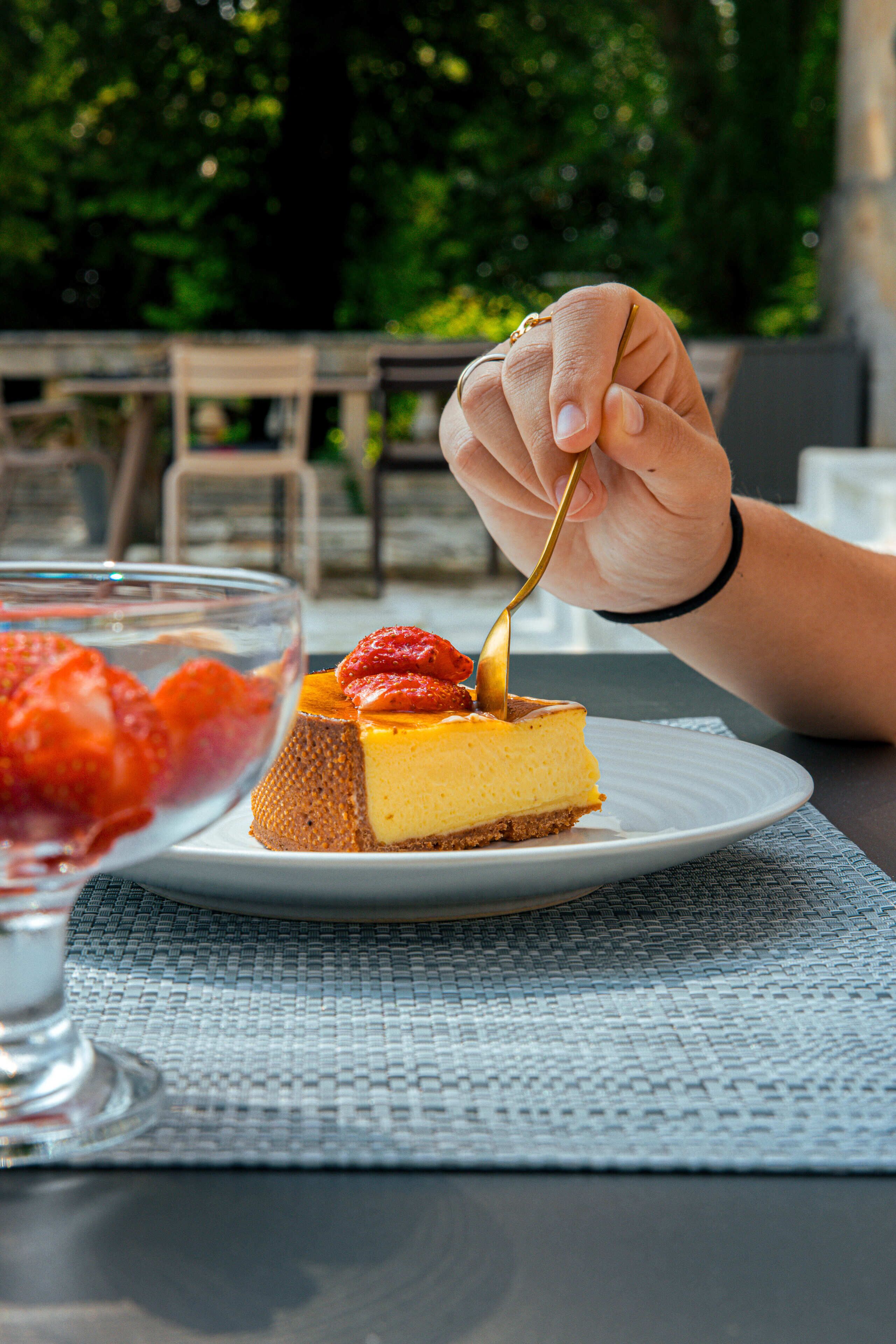 Ein Desserteller mit einem Stück Käsekuchen, belegt mit Erdbeeren, auf einem weißen Unterteller, platziert auf einem Tisch mit einer gewebten Tischmatte.