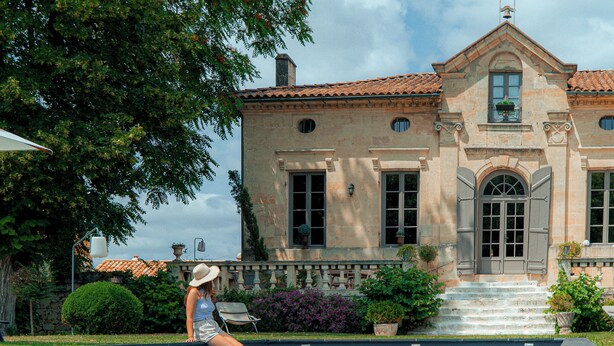 A pool area with a large building in the background, a chair, and a woman sitting by the pool. - Clos Marcamps (Prignac-et-Marcamps)