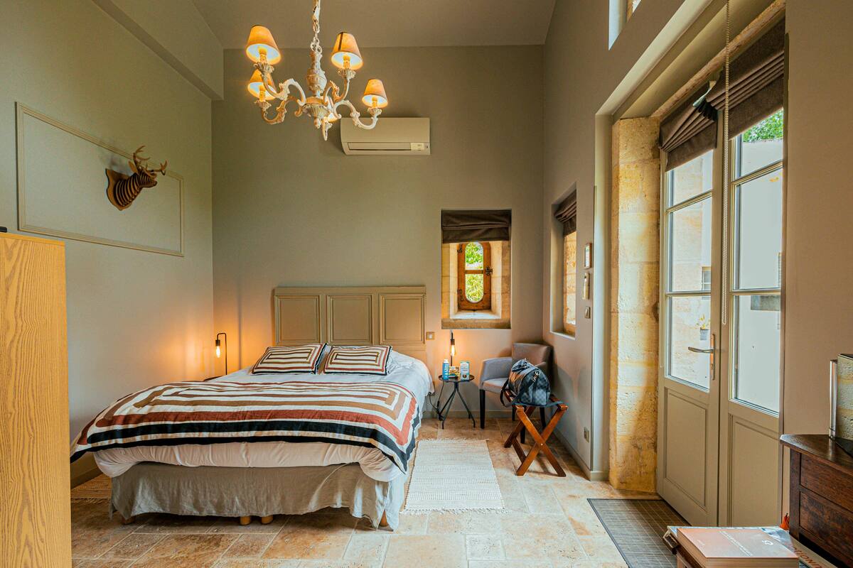 A bedroom with a large bed, a chandelier, a window with curtains, and a stone wall.