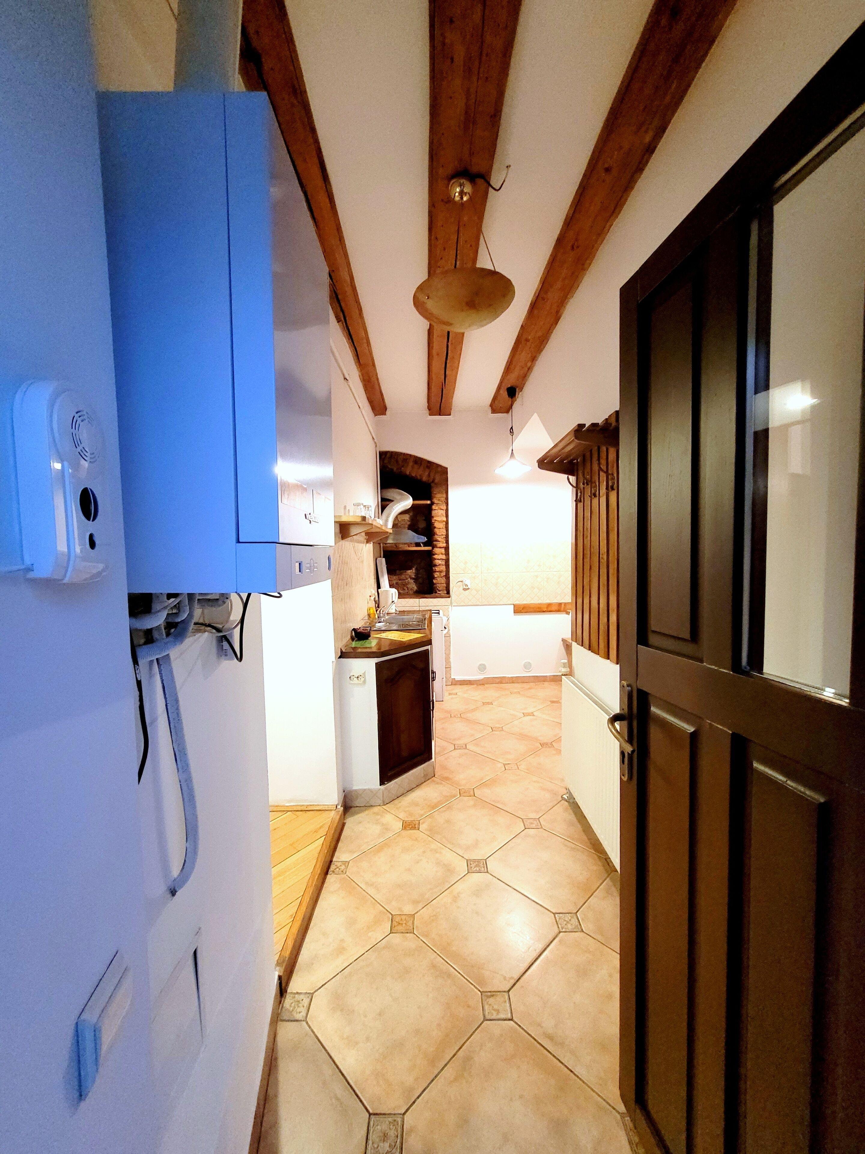 A narrow hallway with a tiled floor, wooden beams on the ceiling, a door on the right, and a small kitchen area with a stove and sink.