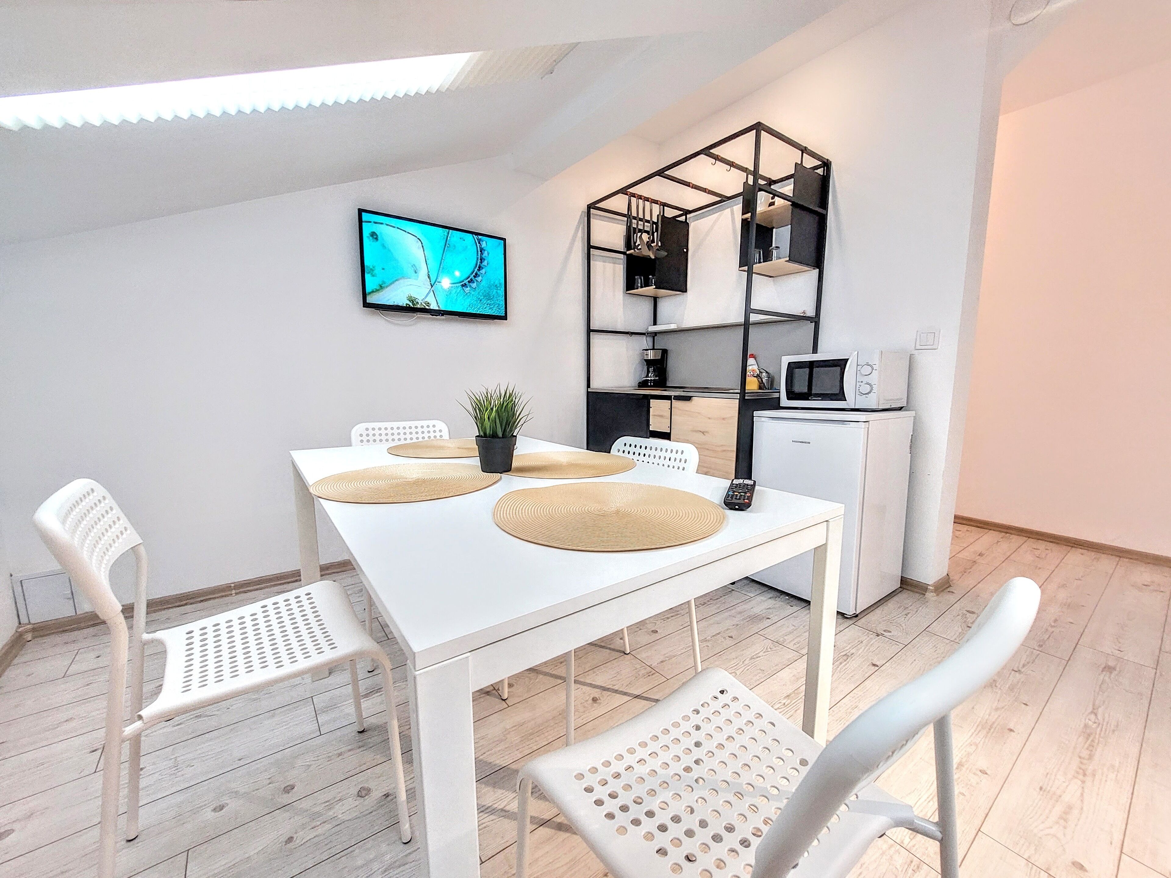 A modern kitchen with white cabinetry, a microwave, and a small refrigerator. There is a dining table with four chairs and a TV mounted on the wall.