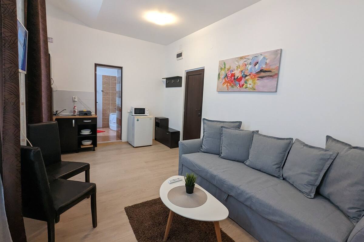 A compact living space with a grey sofa, a small white table, a black chair, a microwave, a refrigerator, and a painting on the wall.