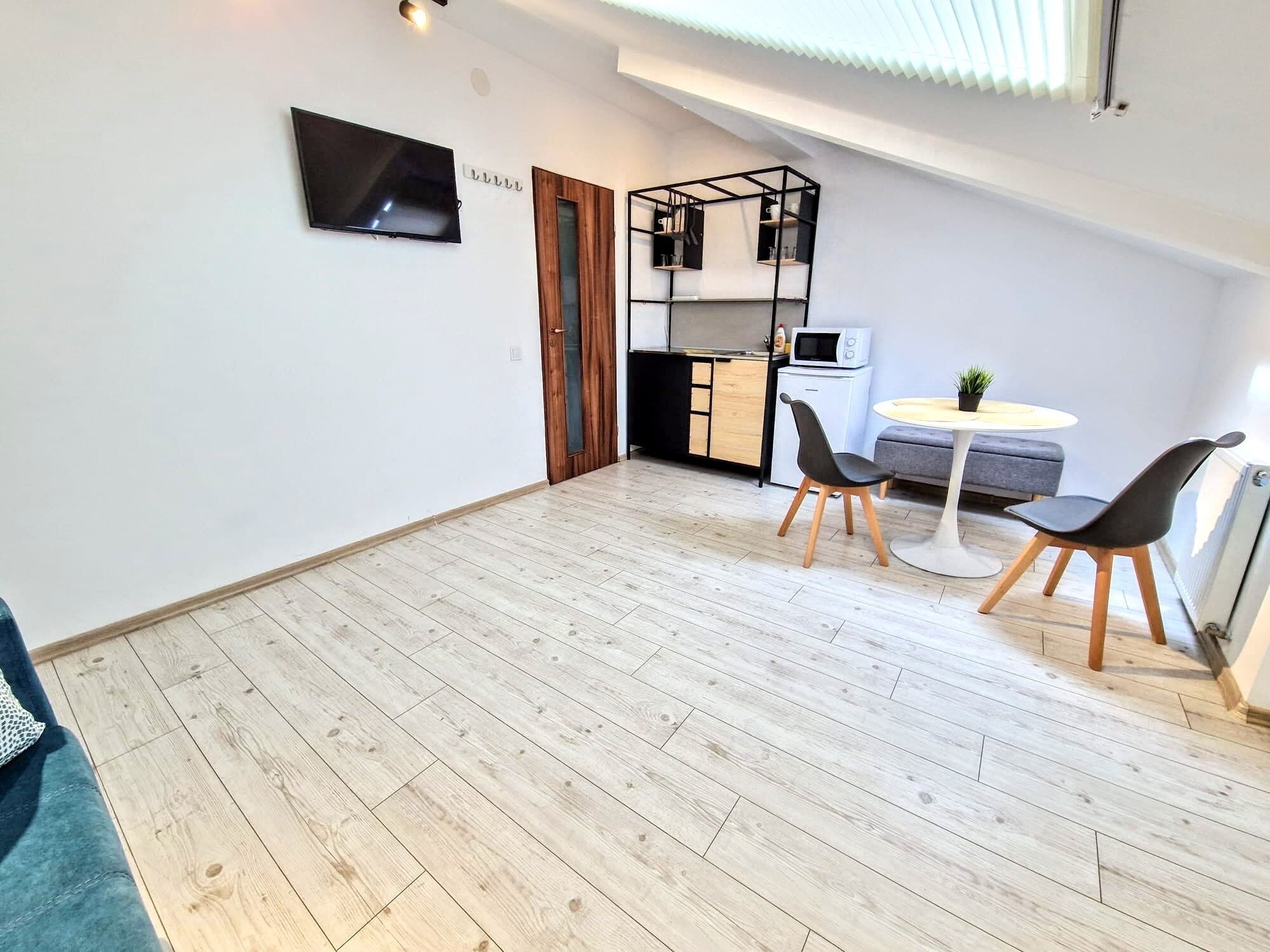 A modern, well-lit room with a wooden floor, a dining table with chairs, a small kitchen area with a microwave, and a TV mounted on the wall.