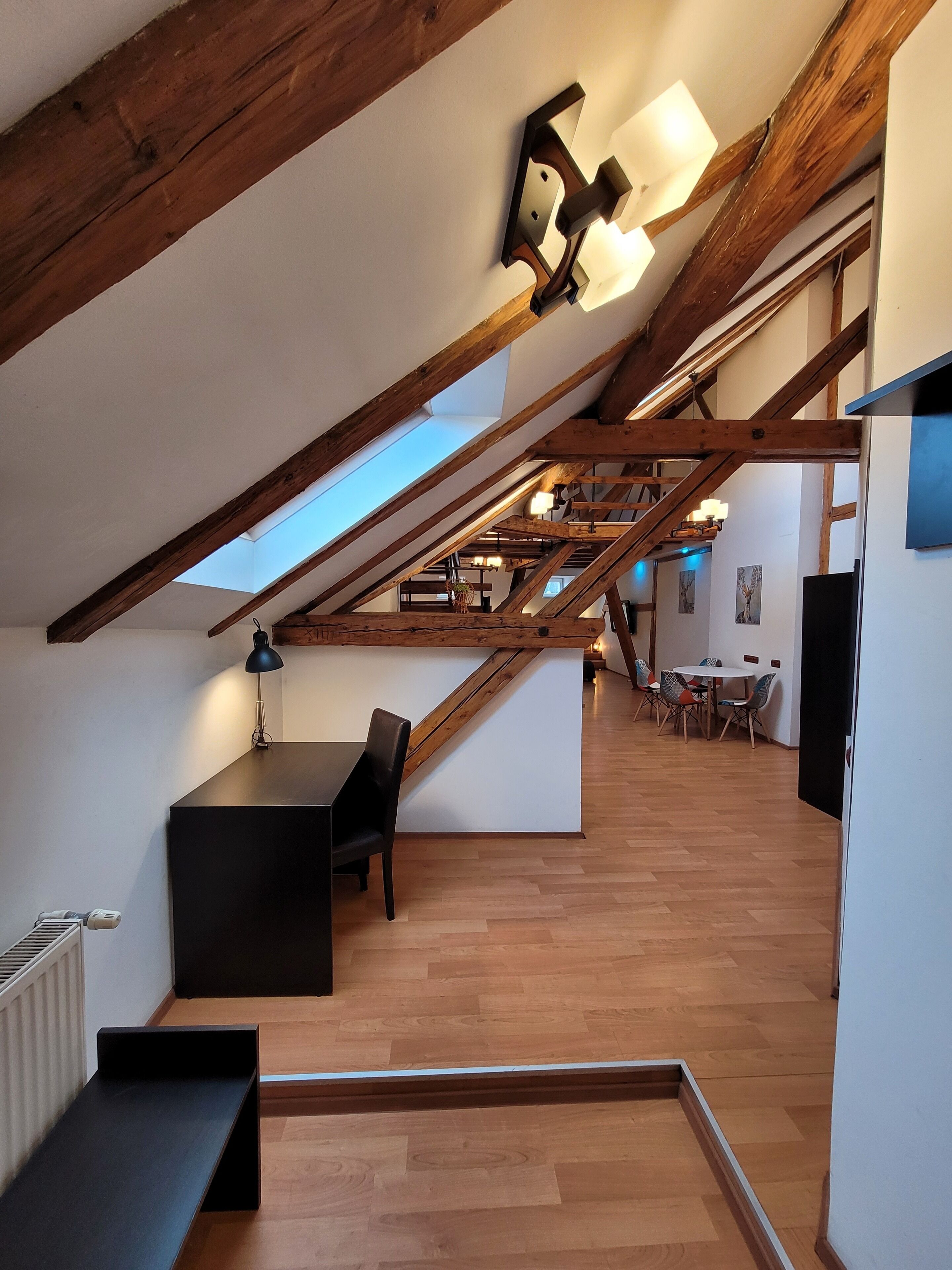 A spacious loft with a sloped ceiling, wooden beams, a skylight, a dining area with chairs, and a black table.