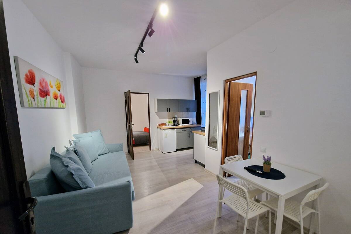 A modern living room with a sofa, a dining table with chairs, a kitchen area, and a door leading to another room.