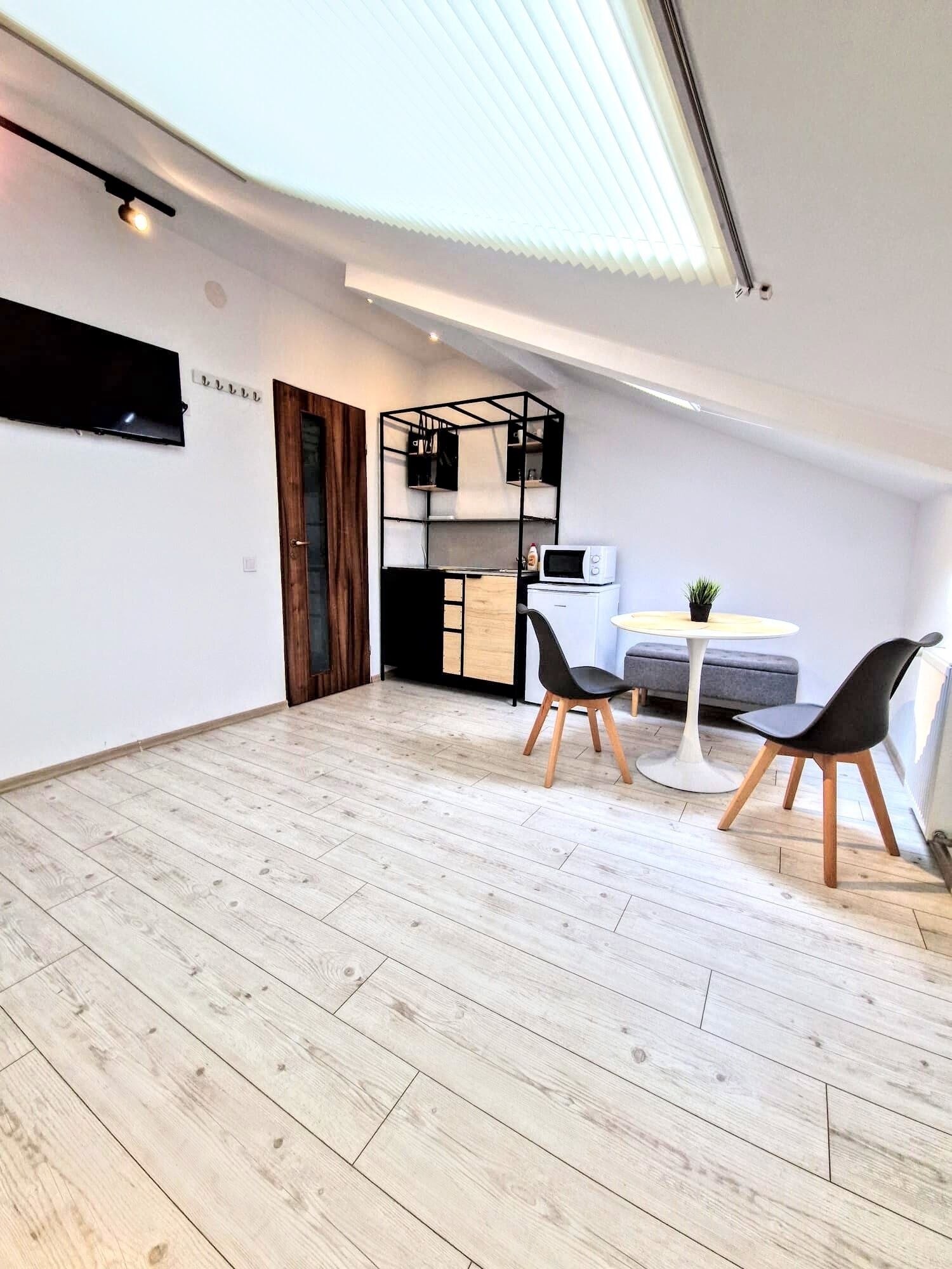Un espacio habitable compacto con cama de altillo, kitchenette, zona de comedor con dos sillas y televisión montada en la pared.