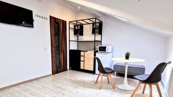 Un espacio habitable compacto con cama de altillo, kitchenette, zona de comedor con dos sillas y televisión montada en la pared.