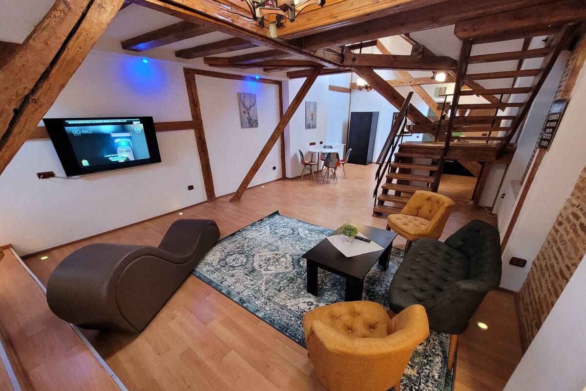 A loft-style living room with exposed wooden beams, a flat-screen TV mounted on the wall, a sofa, two armchairs, a coffee table, and a staircase leading to an upper level.