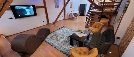 A loft-style living room with exposed wooden beams, a flat-screen TV mounted on the wall, a sofa, two armchairs, a coffee table, and a staircase leading to an upper level.