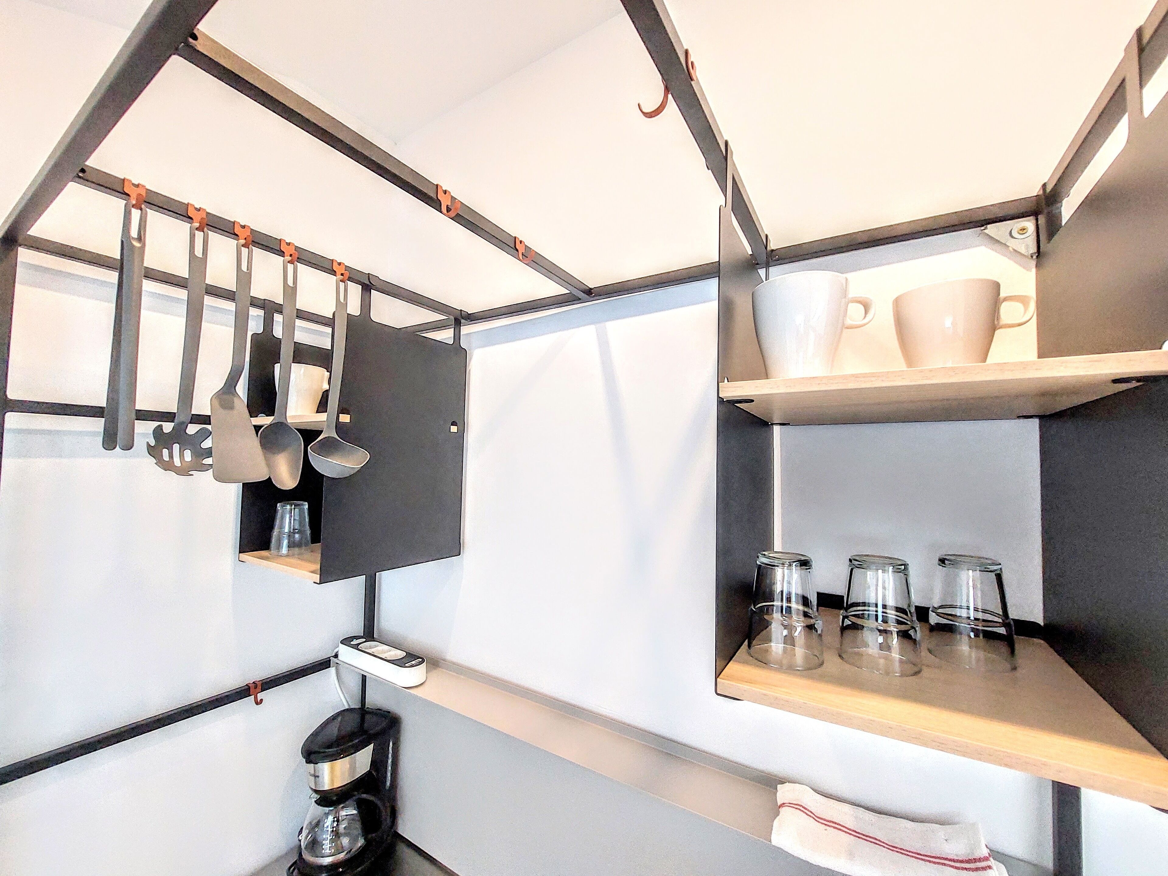Una cuina compacta amb utensilis penjats, una cafetera i prestatges amb gots i tasses.