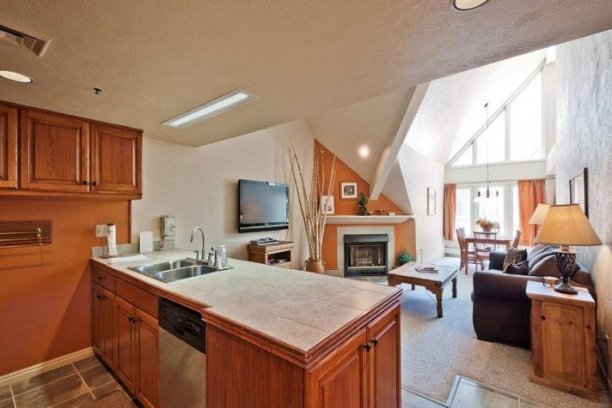 A spacious kitchen with wooden cabinets, a built-in dishwasher, and a large island. There is a fireplace, a dining area with a table and chairs, and a living room with a sofa and a lamp.