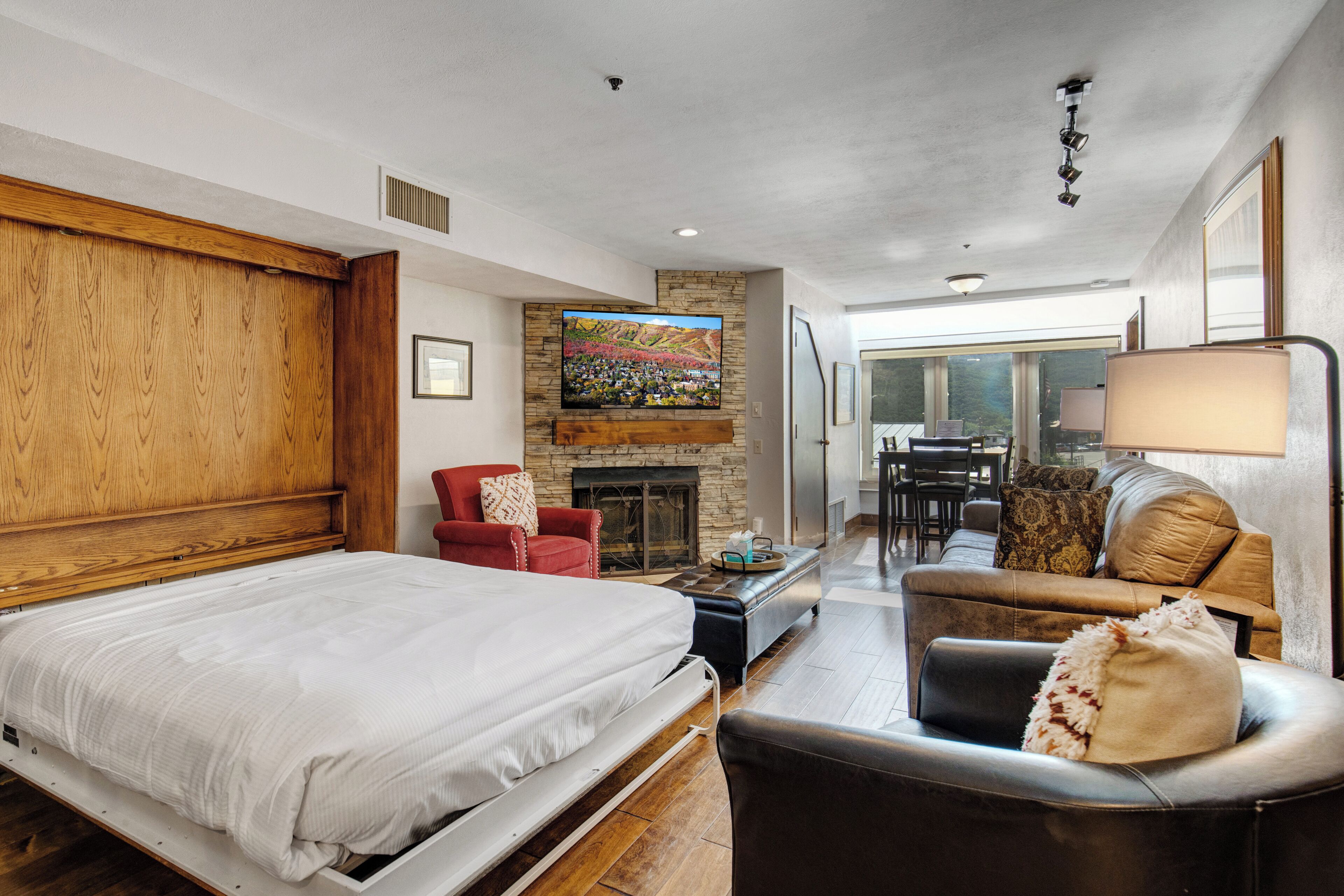 Quarto moderno com uma cama grande, uma poltrona vermelha, lareira de pedra e uma área de jantar visível por uma porta de vidro.