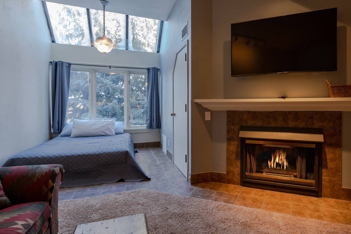 A bedroom with a bed, a fireplace, a TV mounted above it, and a window with curtains.
