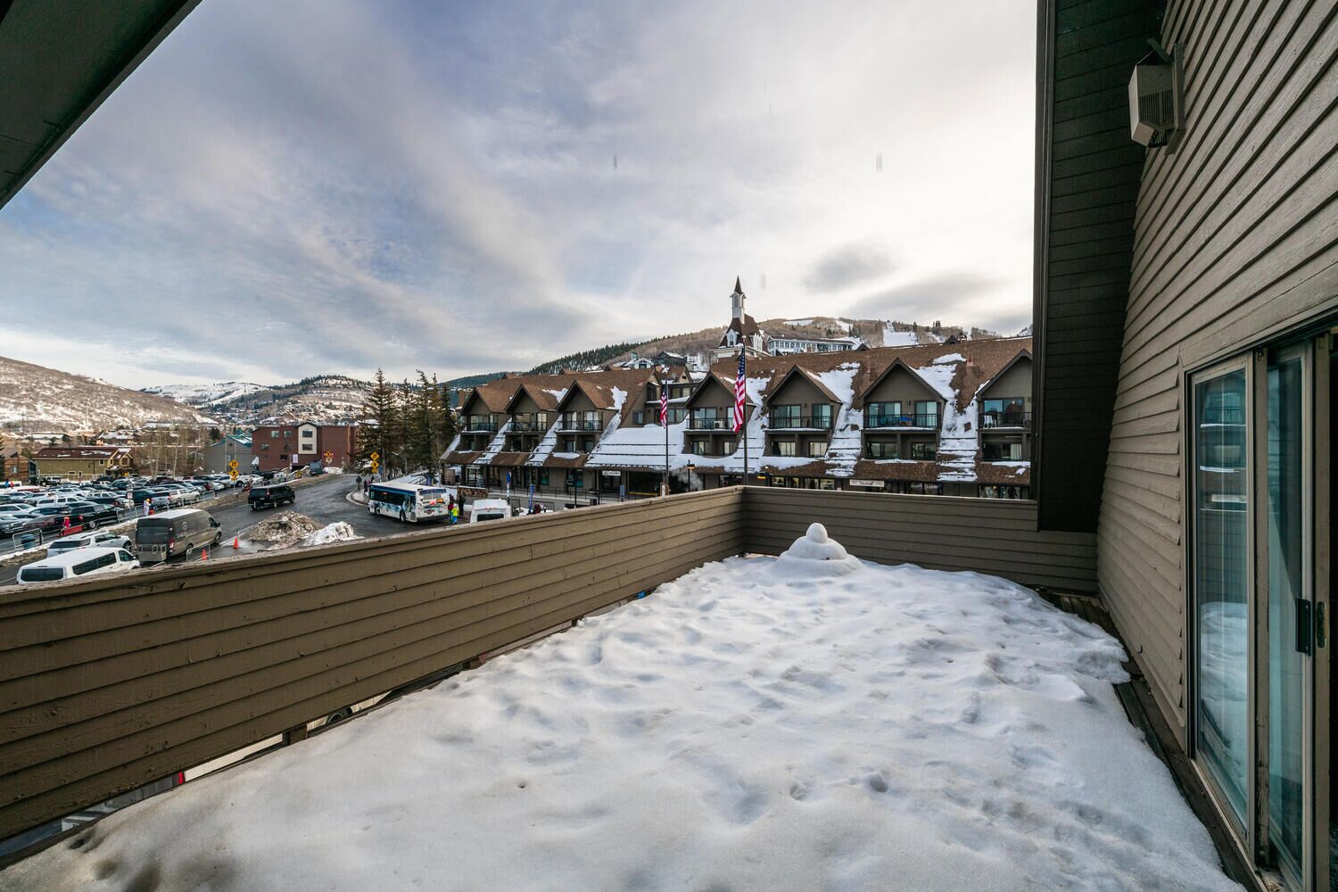 A hotel with a snow-covered balcony, a parking lot, and a view of a town with buildings and a mountain in the background.