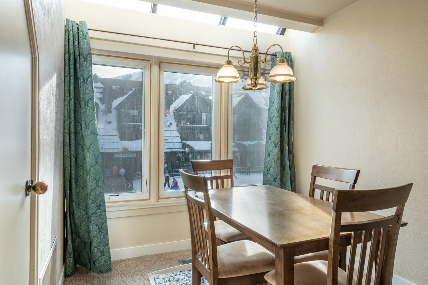 A dining area with a wooden table and chairs, a chandelier, and a view of snowy mountains through large windows.