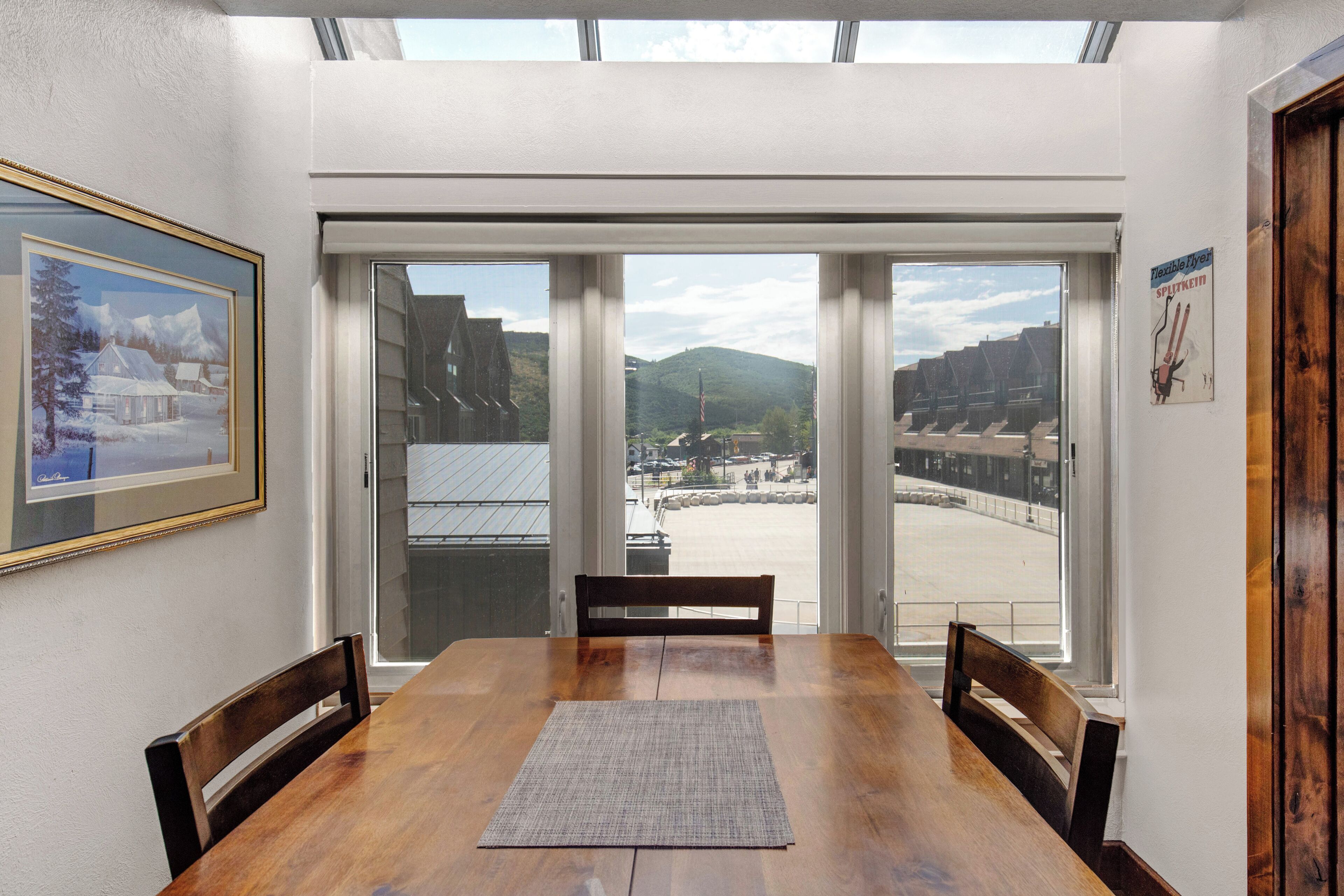 Uma área de refeições com uma mesa e cadeiras de madeira, janelas amplas com vista para uma paisagem nevada e edifícios, e um quadro na parede.