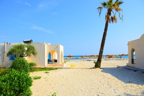 A beach with a palm tree, a building with arches, and beach umbrellas in the background. - Royal Lido Resort & Spa (Dar Chaabane Al Fehri)
