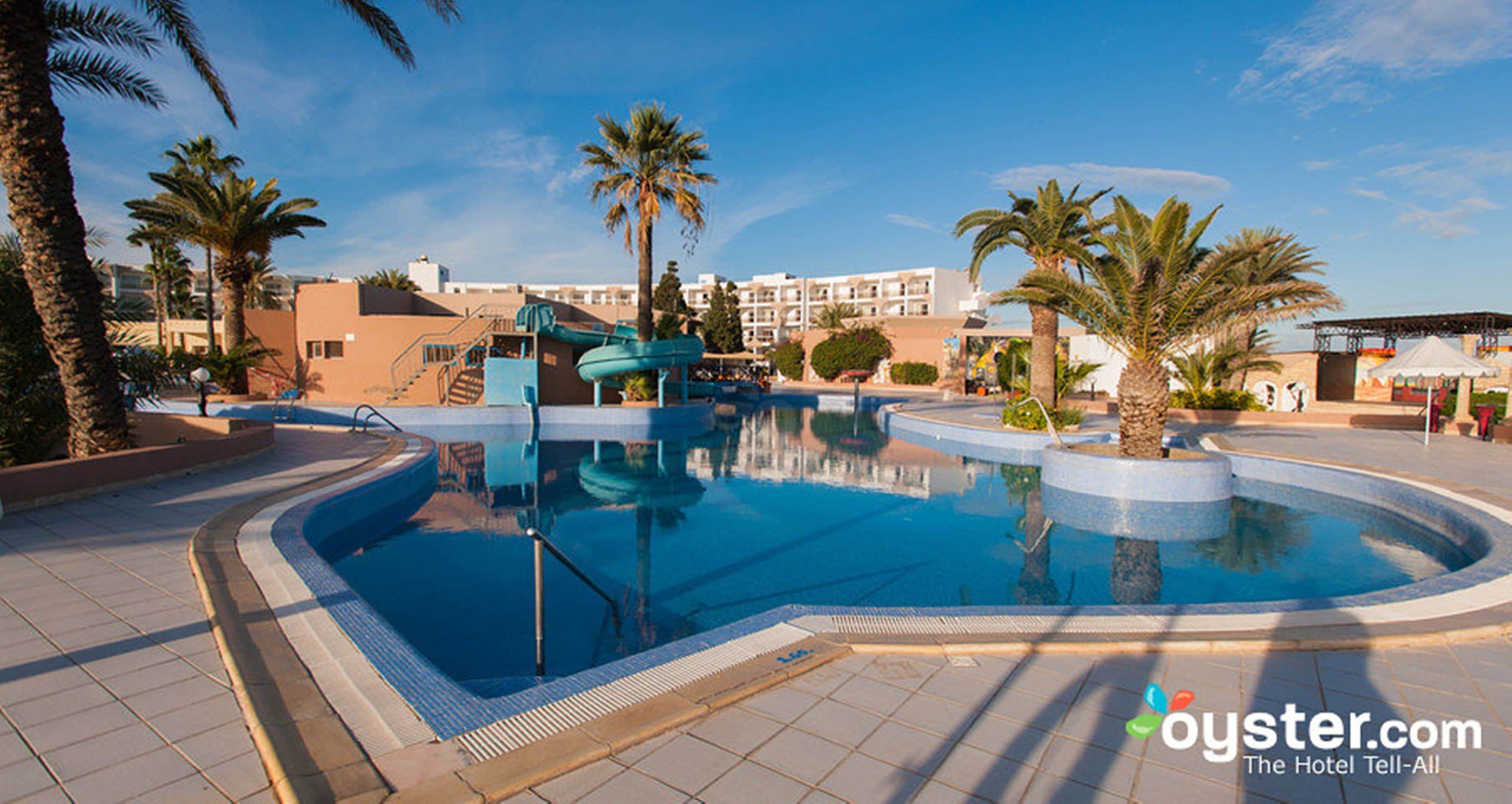 Une piscine avec un toboggan, des palmiers et un bâtiment d’hôtel en arrière-plan.