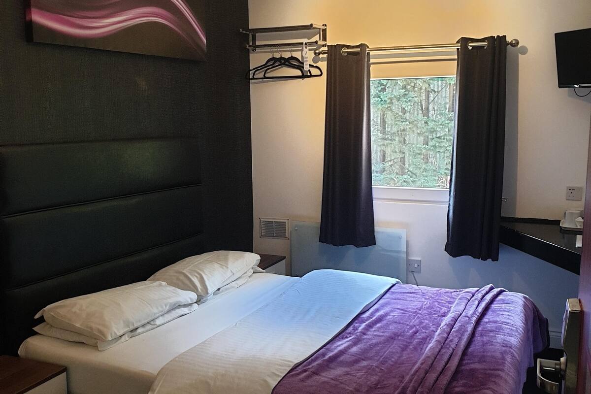 A hotel room with a bed, a window with curtains, a TV mounted on the wall, and a coat rack.