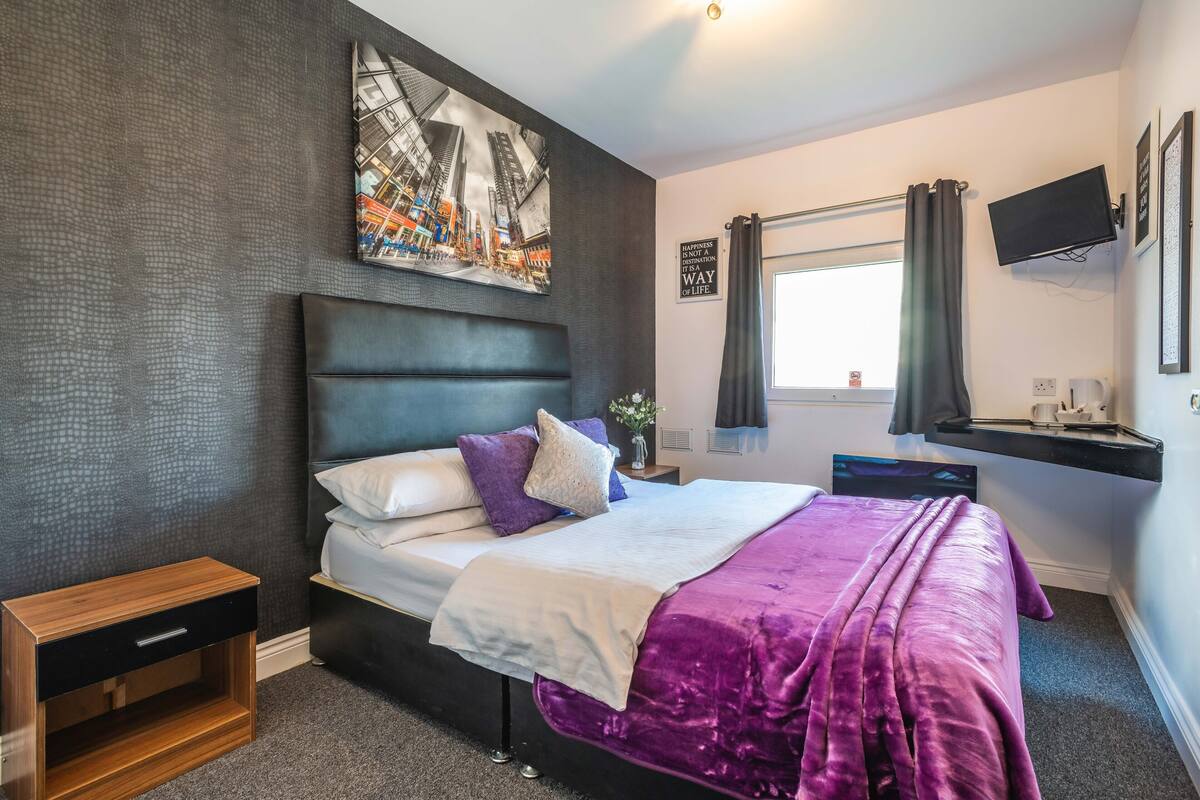 A modern bedroom with a large bed, a nightstand, a flat-screen TV, and a window with curtains.