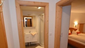 A hotel room with a wooden door, a bathroom with a shower, and a bed with pillows. - Haus Almfriede (Leutasch)
