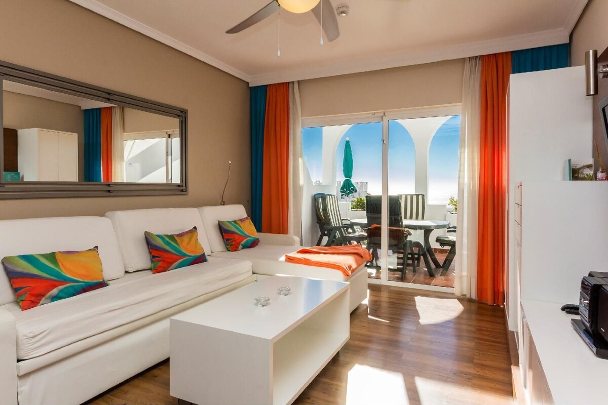 A modern living room with a white sofa, colorful pillows, a coffee table, and a balcony with outdoor furniture.
