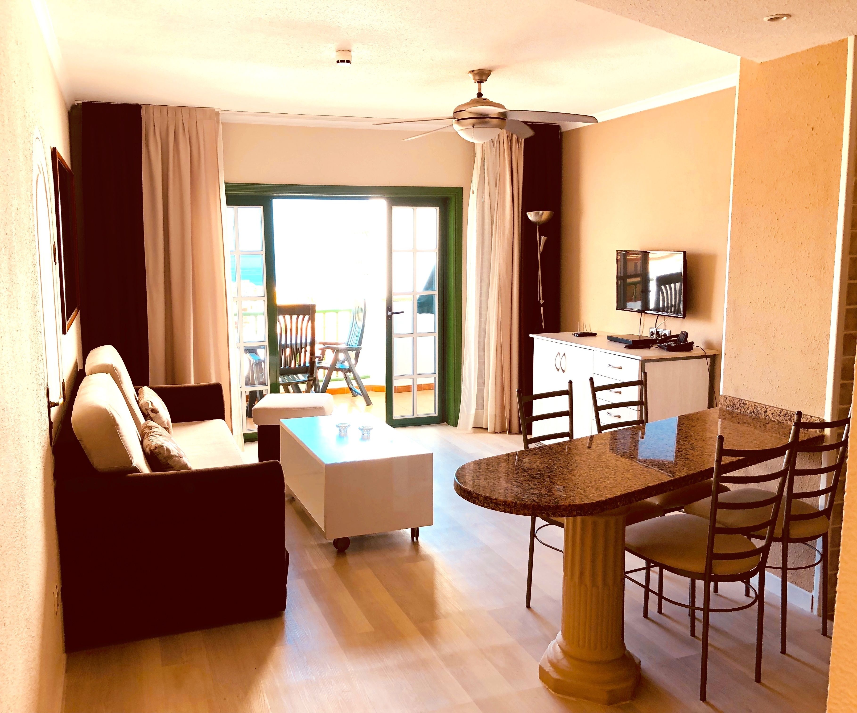 A hotel room with a living area, dining table, chairs, a sofa, a TV, and a balcony with chairs.