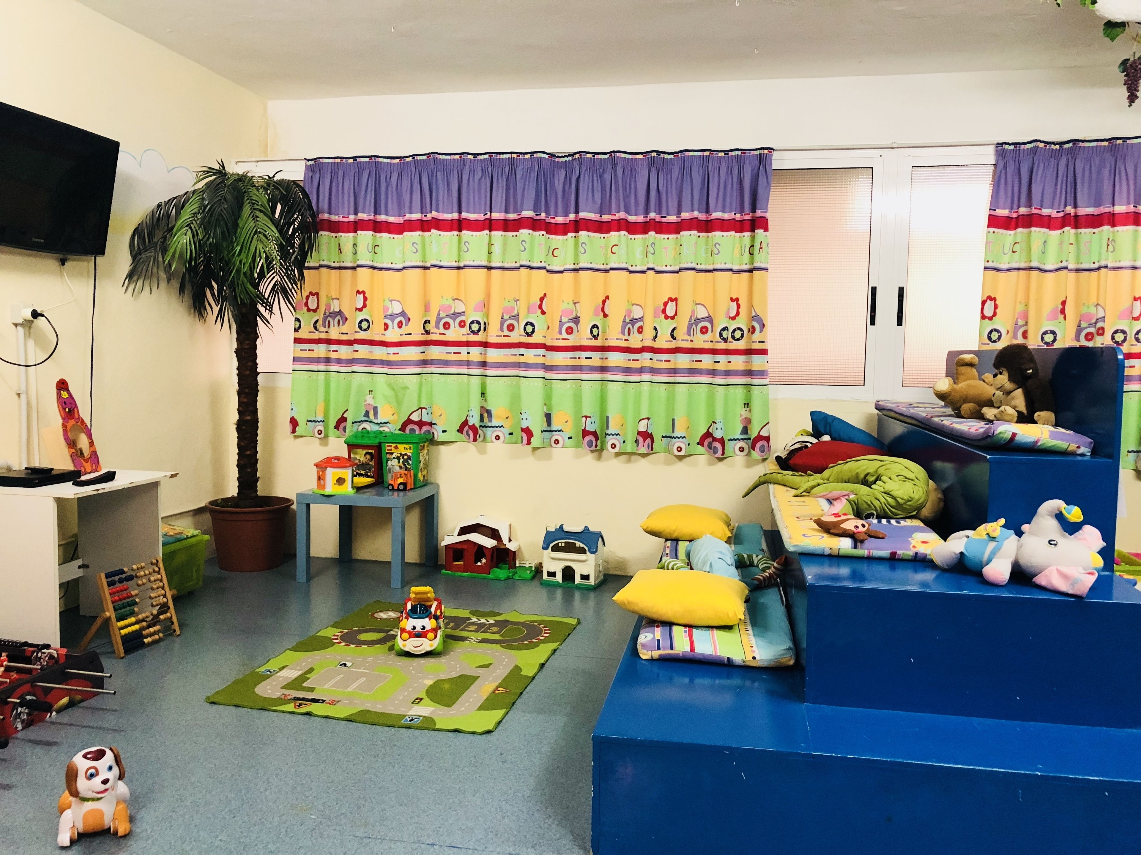 Un cuarto de juegos para niños con un televisor grande, cortinas coloridas, un tapete de juego, juguetes y una estructura de juego azul.