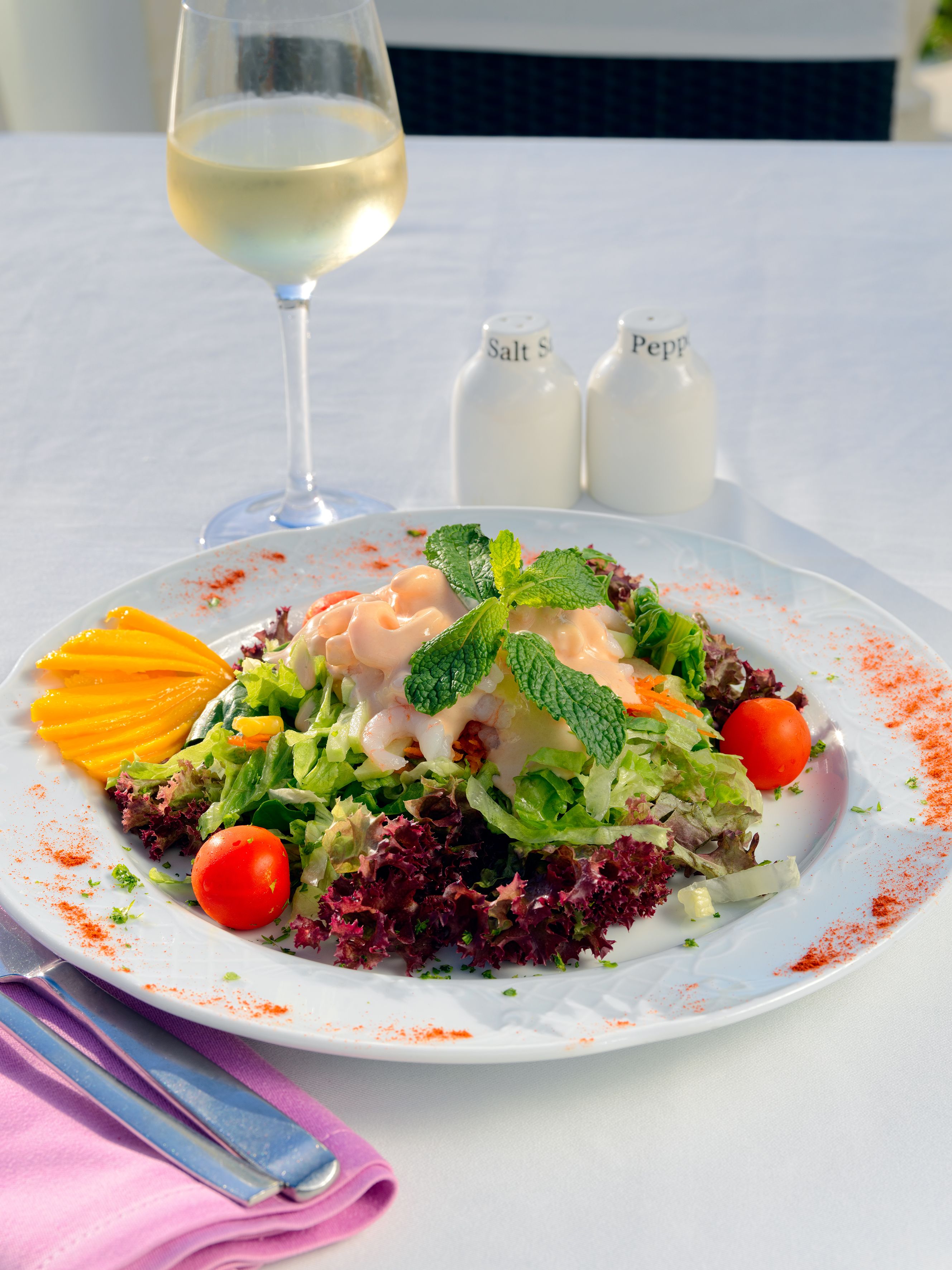Un plato blanco con ensalada, una copa de vino blanco y saleros y pimenteros sobre una mesa.