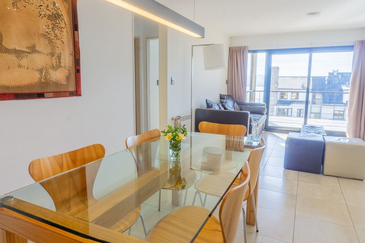 Un salon moderne avec une table à manger en verre, des chaises en bois, un canapé et une vue sur la ville grâce à de grandes fenêtres.