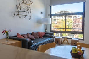 Una sala moderna con un sofá de cuero negro, una mesa de comedor de madera con un frutero, y un ventanal que ofrece vistas a un edificio y árboles.
