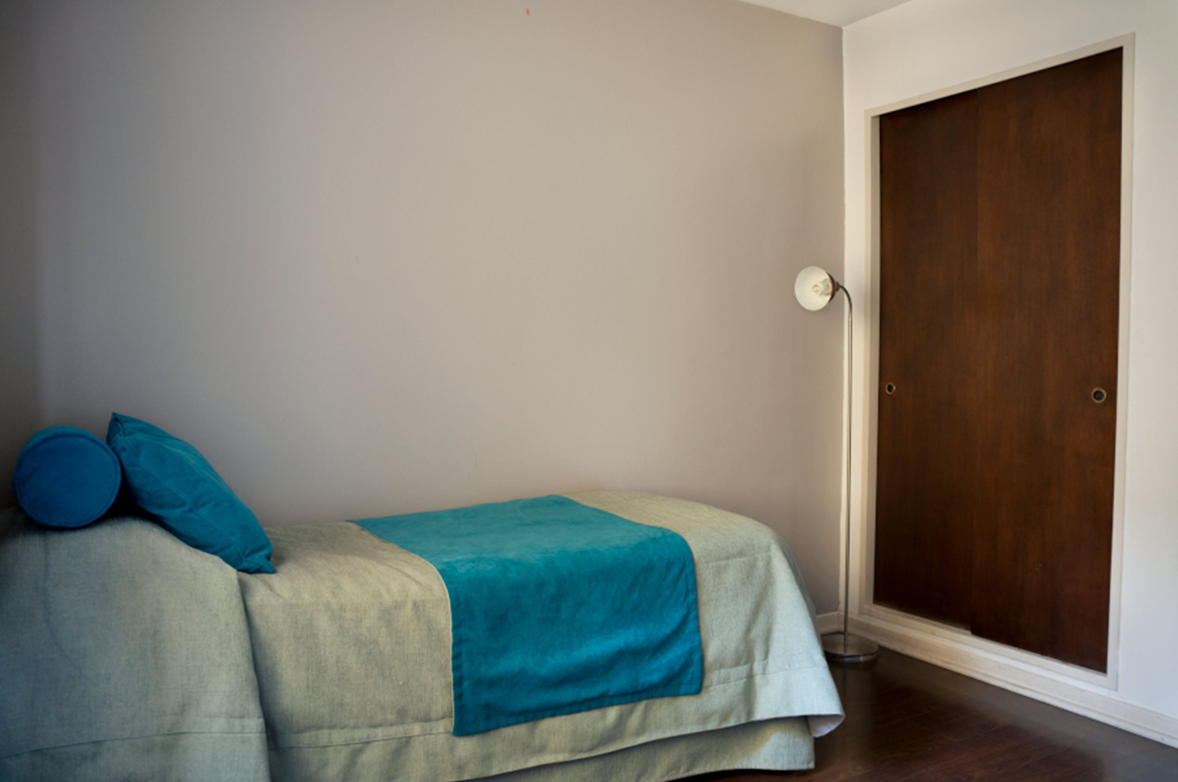 Uma cama pequena e bem arrumada, com um cobertor e travesseiros azul-esverdeados, uma porta de madeira e uma parede lisa.