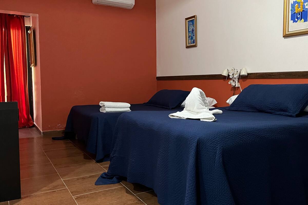 Hotellihuoneessa on kaksi sänkyä, joissa on sininen peitto ja valkoiset tyynyt. Vasemmalla puolella on punainen verho, seinällä on kehystetty kuva ja huoneessa on pieni pöytä, jonka päällä on siististi taiteltuja pyyhkeitä.